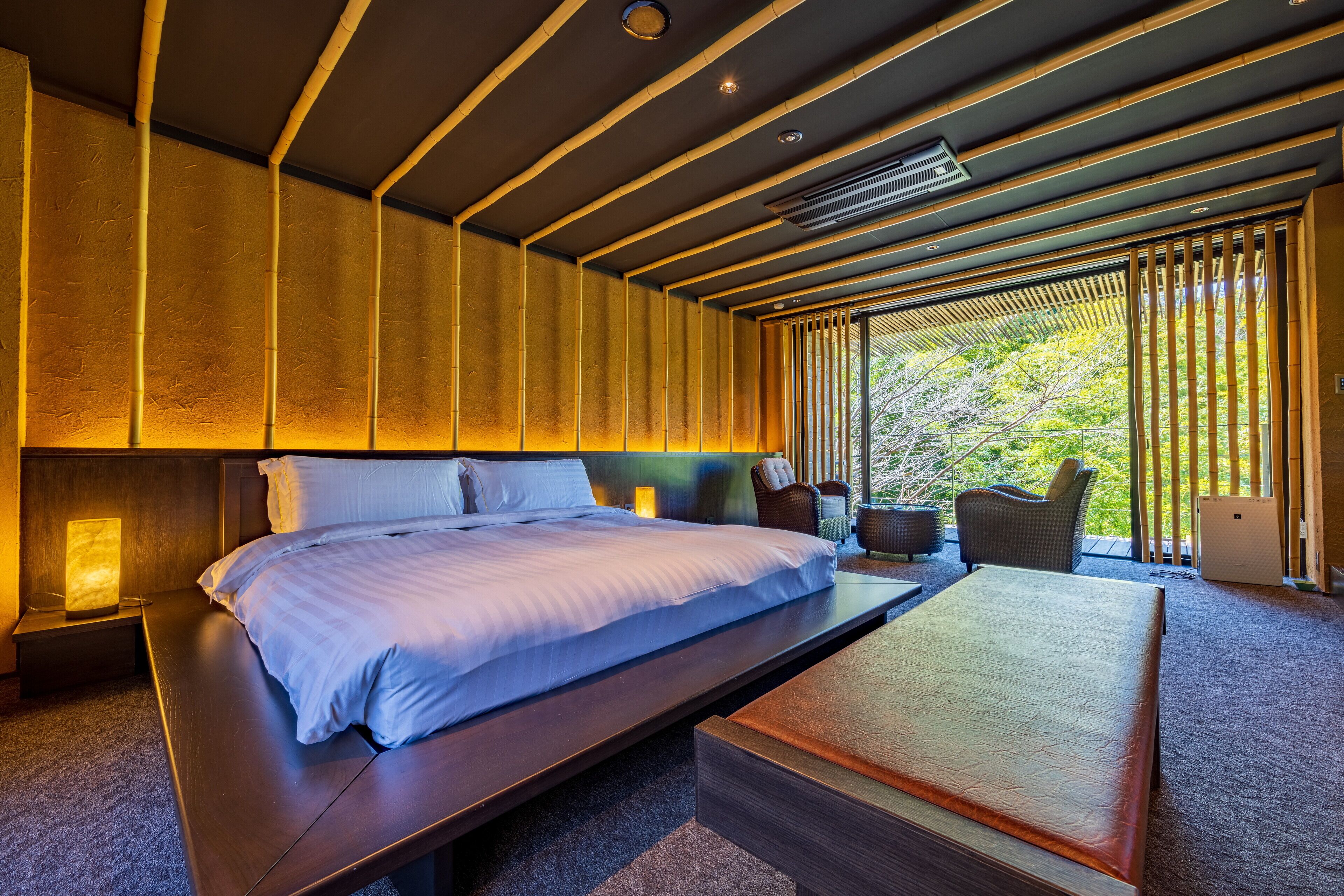 A modern hotel room with a large bed, bedside tables, a bedside lamp, a sofa, and a view of greenery through the window.