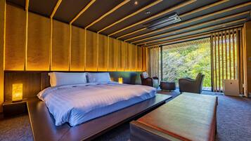 A modern hotel room with a large bed, bedside tables, a bedside lamp, a sofa, and a view of greenery through the window.