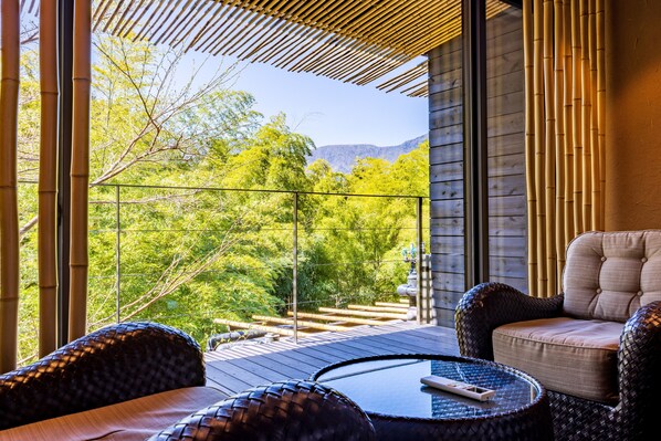 A balcony with wicker furniture, a glass railing, and a view of trees and mountains.
