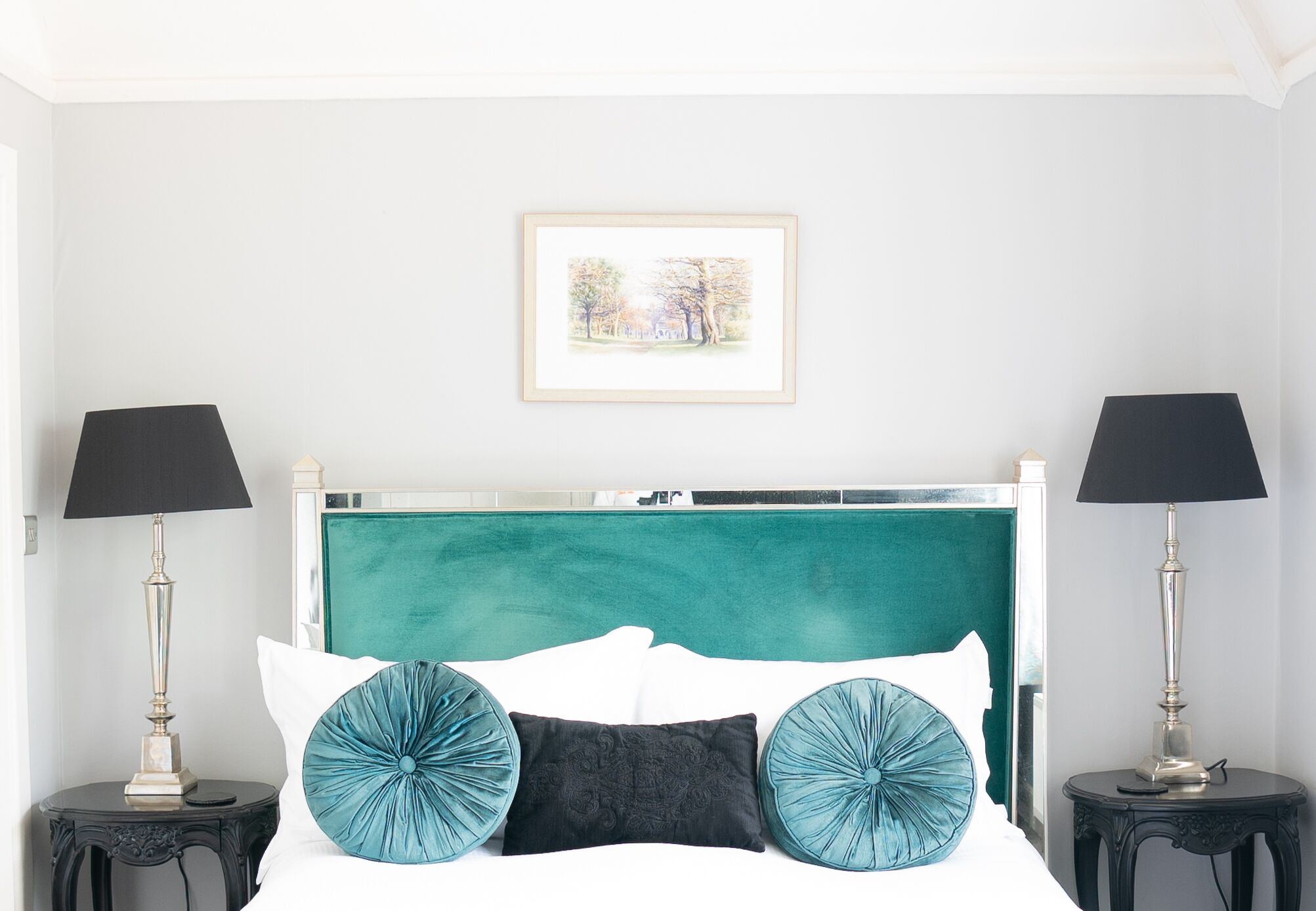 Un dormitorio ordenado con cabecera color turquesa, ropa de cama blanca y dos lámparas negras sobre mesitas de noche.