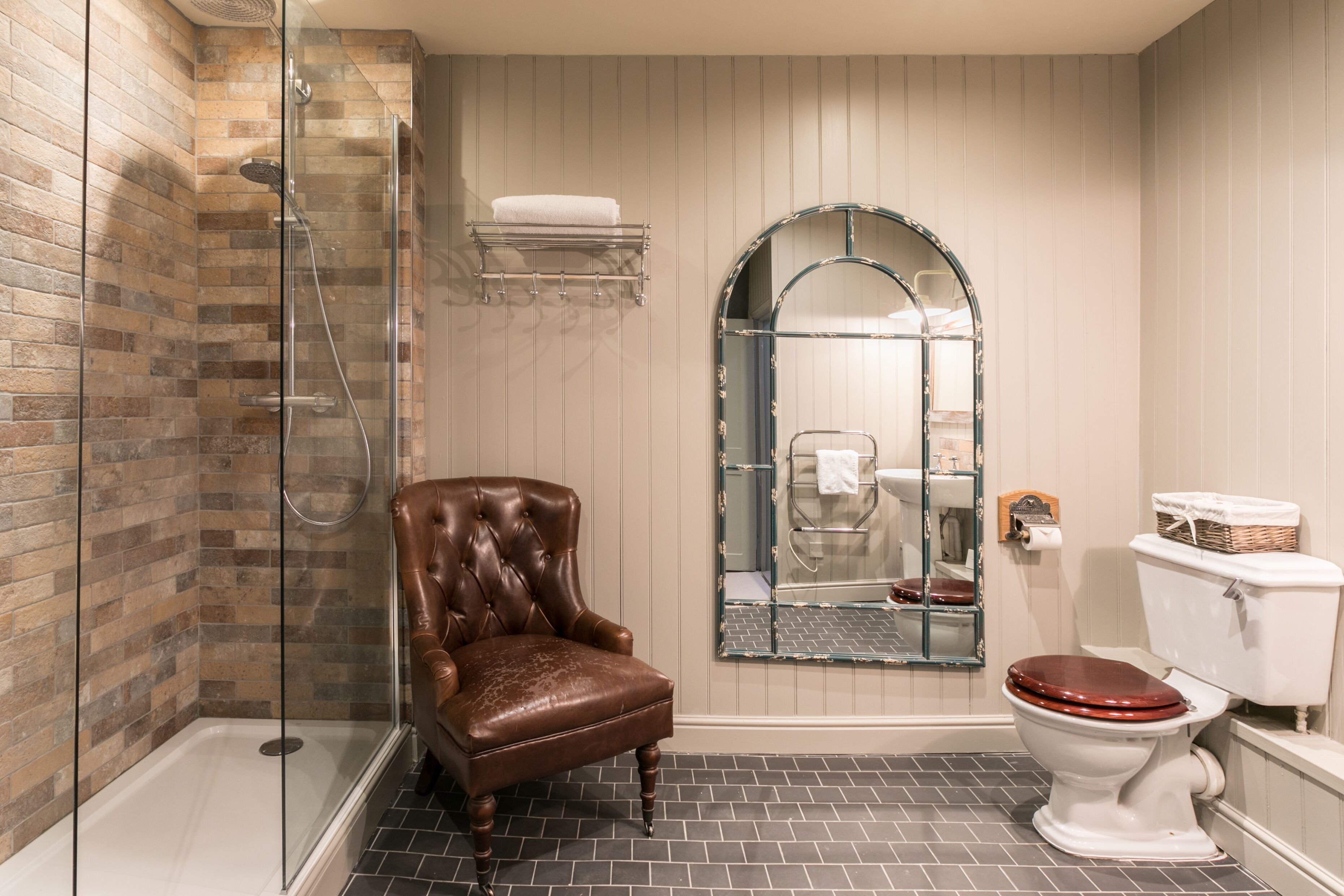 A bathroom with a glass shower enclosure, a brown leather chair, a white toilet, and a mirror with an arched frame.