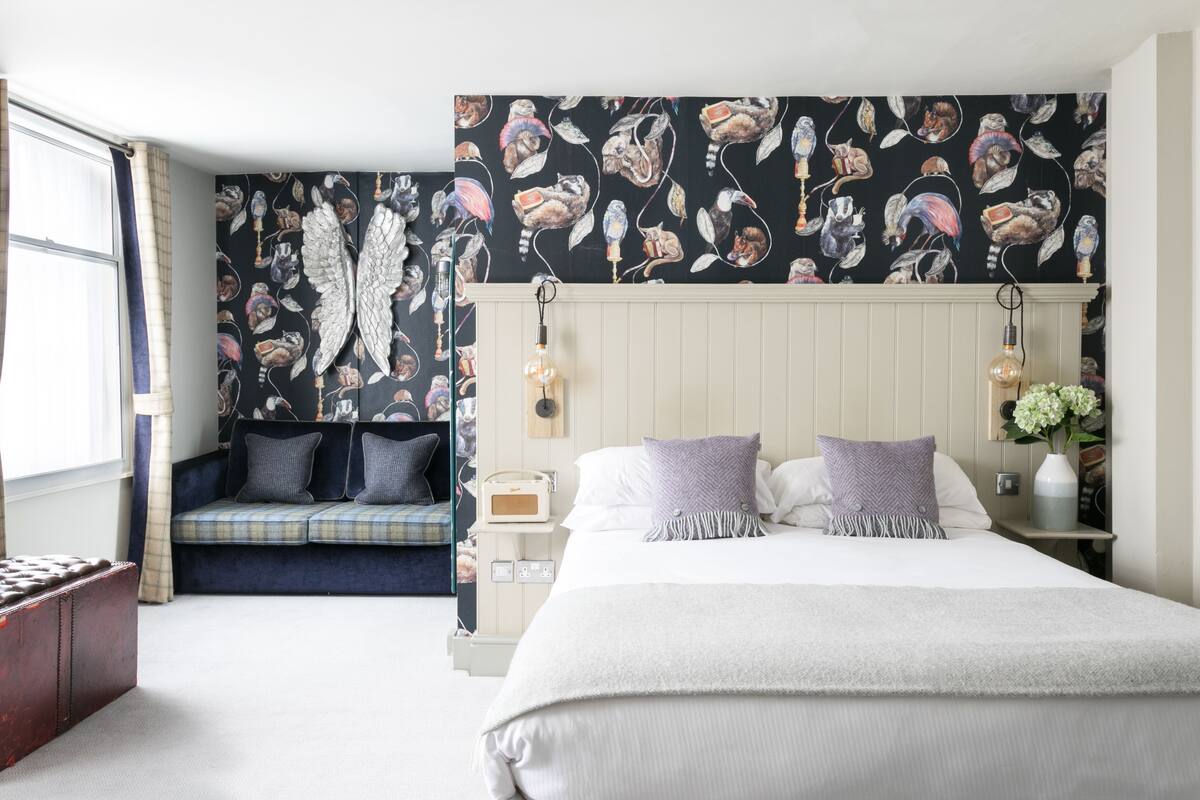 A bedroom with a large bed, a bench with cushions, a wall with a cat pattern, and a window with curtains.