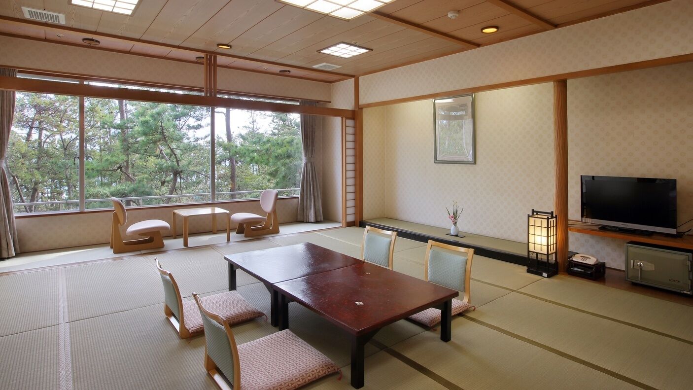 Ein traditioneller japanischer Raum mit Tatami-Böden, einem niedrigen Tisch, Stühlen, einem Fernseher und Blick auf Bäume.