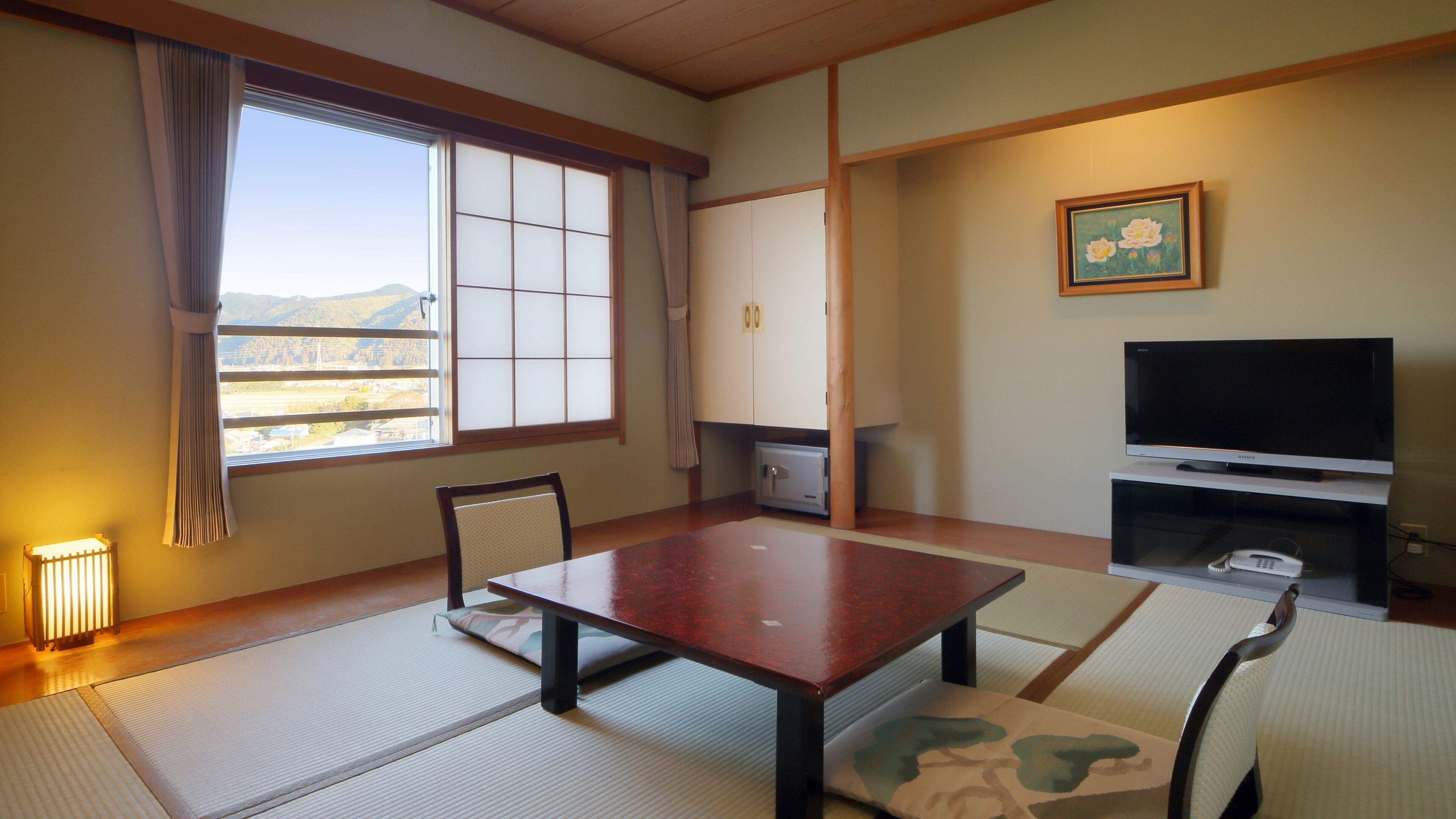 A room with tatami mats, a low table, chairs, a TV, a cabinet, and a window with a view of hills.