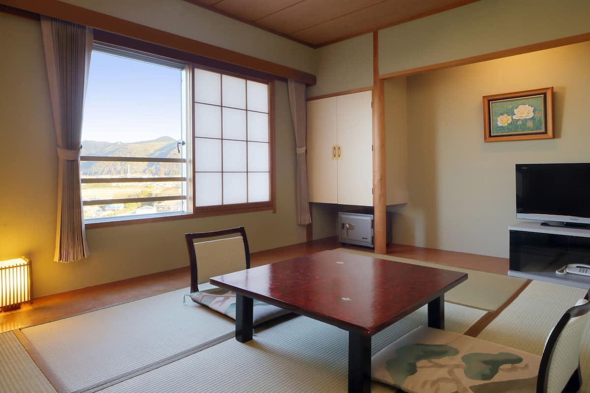 A room with tatami mats, a low table, chairs, a TV, a cabinet, and a window with a view of hills.