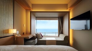 A hotel room with a bed, a desk with a lamp, a TV mounted on the wall, and a window with a view of the sea. - Kamogawakan (Kamogawa)