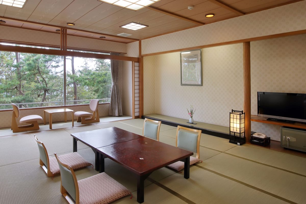 A traditional Japanese-style room with tatami flooring, a low table, chairs, a television, and a view of trees outside.