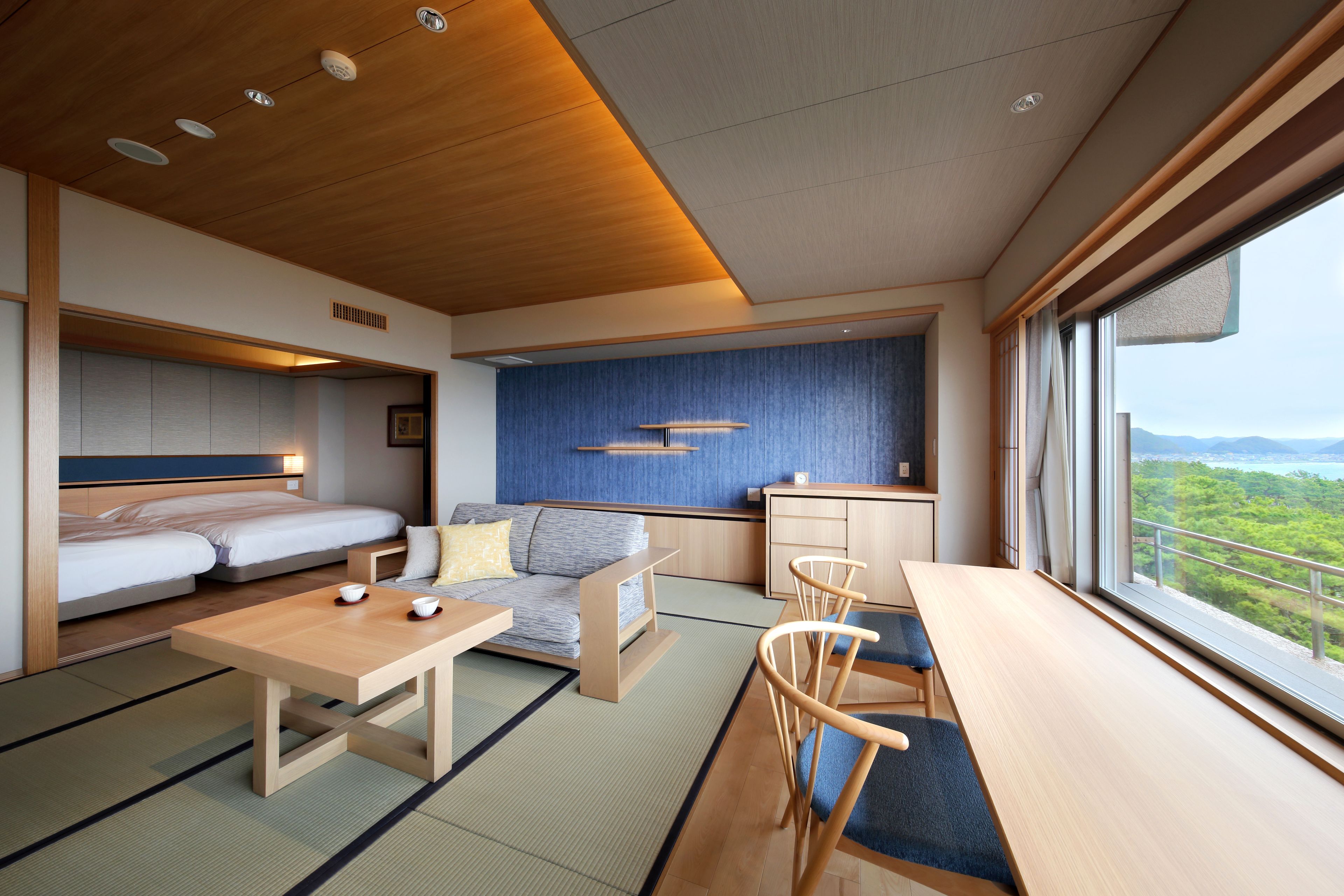 Habitación de hotel moderna con cama, sofá, mesa de comedor y sillas. Cuenta con una zona de cocina con fregadero y armarios. La habitación tiene ventanas amplias con vistas a zonas verdes y montañas.