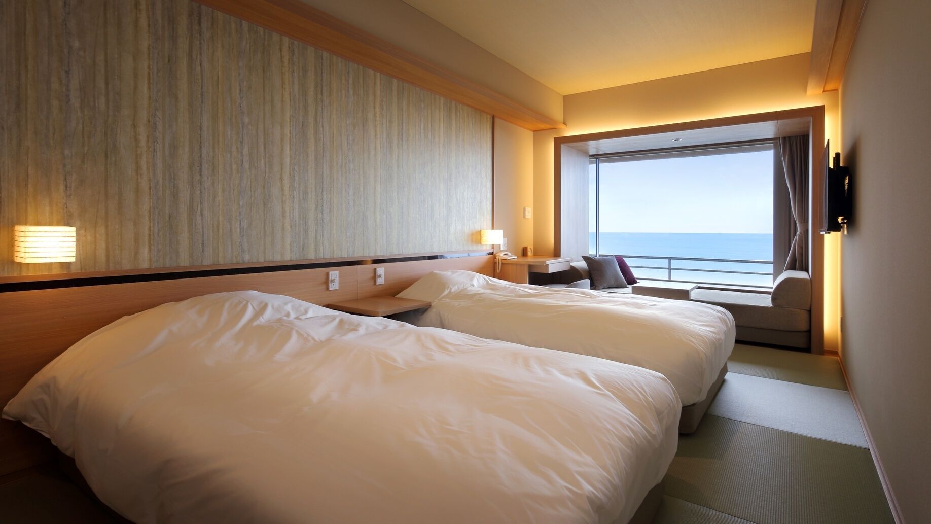 Ein Hotelzimmer mit zwei Betten, einem Schreibtisch, einem Stuhl, einem großen Fenster mit Meerblick und einem an der Wand befestigten Fernseher.