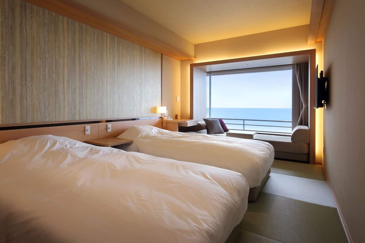 A hotel room with two beds, a desk, a chair, a large window with a view of the sea, and a wall-mounted TV.