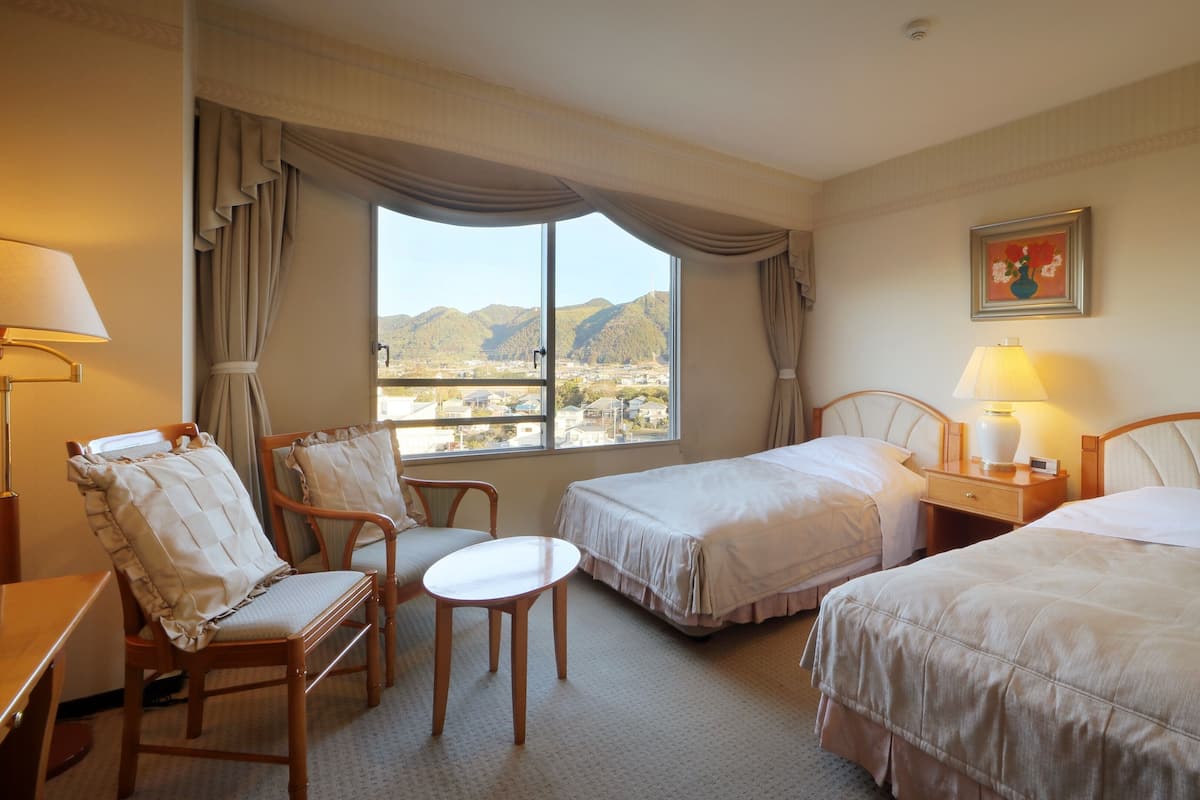 A hotel room with two beds, a chair, a small table, and a lamp.