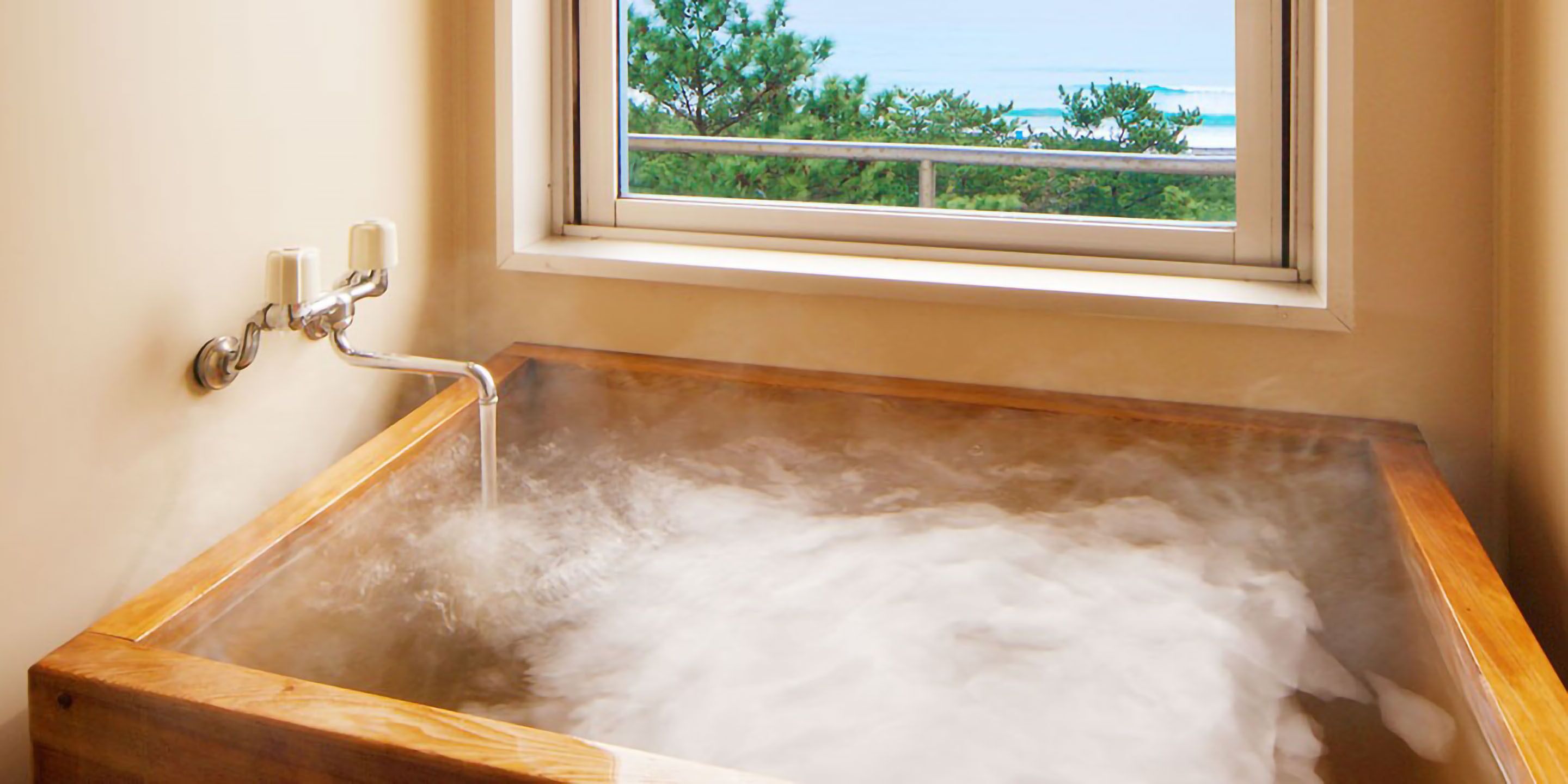 A wooden bathtub filled with water, a window with a view of trees and a body of water, and a wall-mounted faucet.