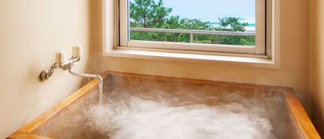 A wooden bathtub filled with water, a window with a view of trees and a body of water, and a wall-mounted faucet.