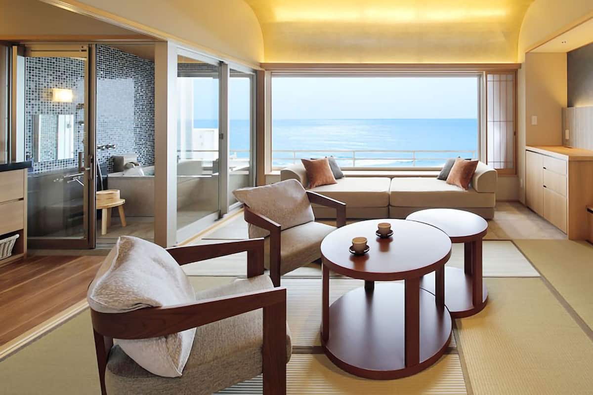 A modern living room with a large TV, comfortable seating, and a view of the sea.