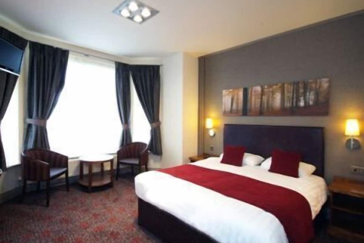 A hotel room with a large bed, two chairs, a desk, and a window with curtains.