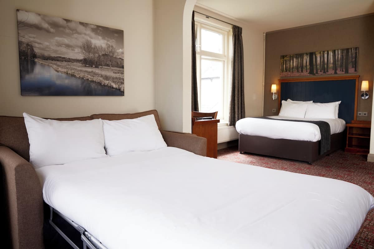 A hotel room with a large bed, a smaller bed, a sofa, a window with curtains, and a painting on the wall.