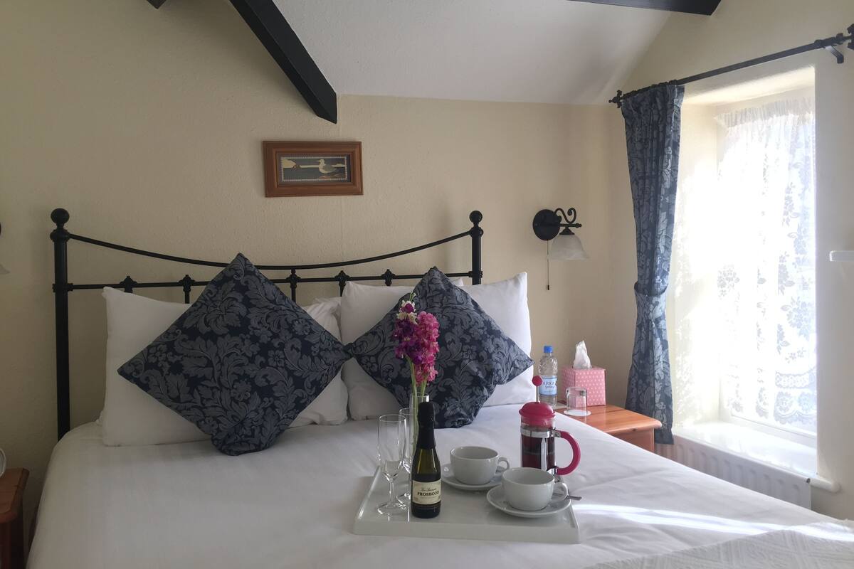 A neatly made bed with patterned pillows, a bedside table with a bottle and glasses, and a window with curtains.