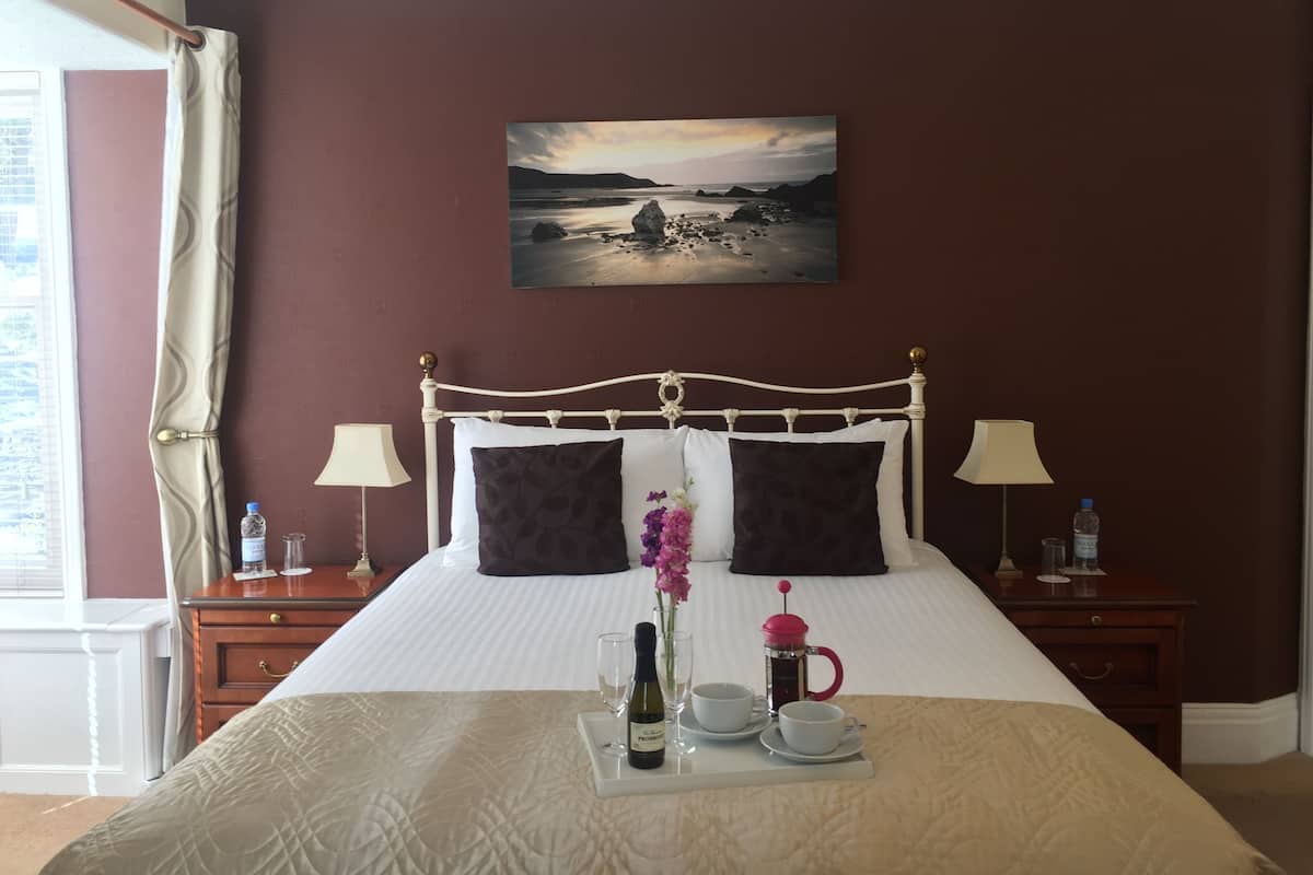 A neatly made bed with a tray of breakfast items, a painting of a coastal scene, and a view of the outdoors through a window.