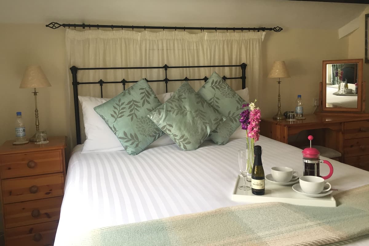 A neatly made bed with floral pillows, a bedside table with a lamp, a water bottle, and a tray with a bottle of wine, cups, and a thermos.