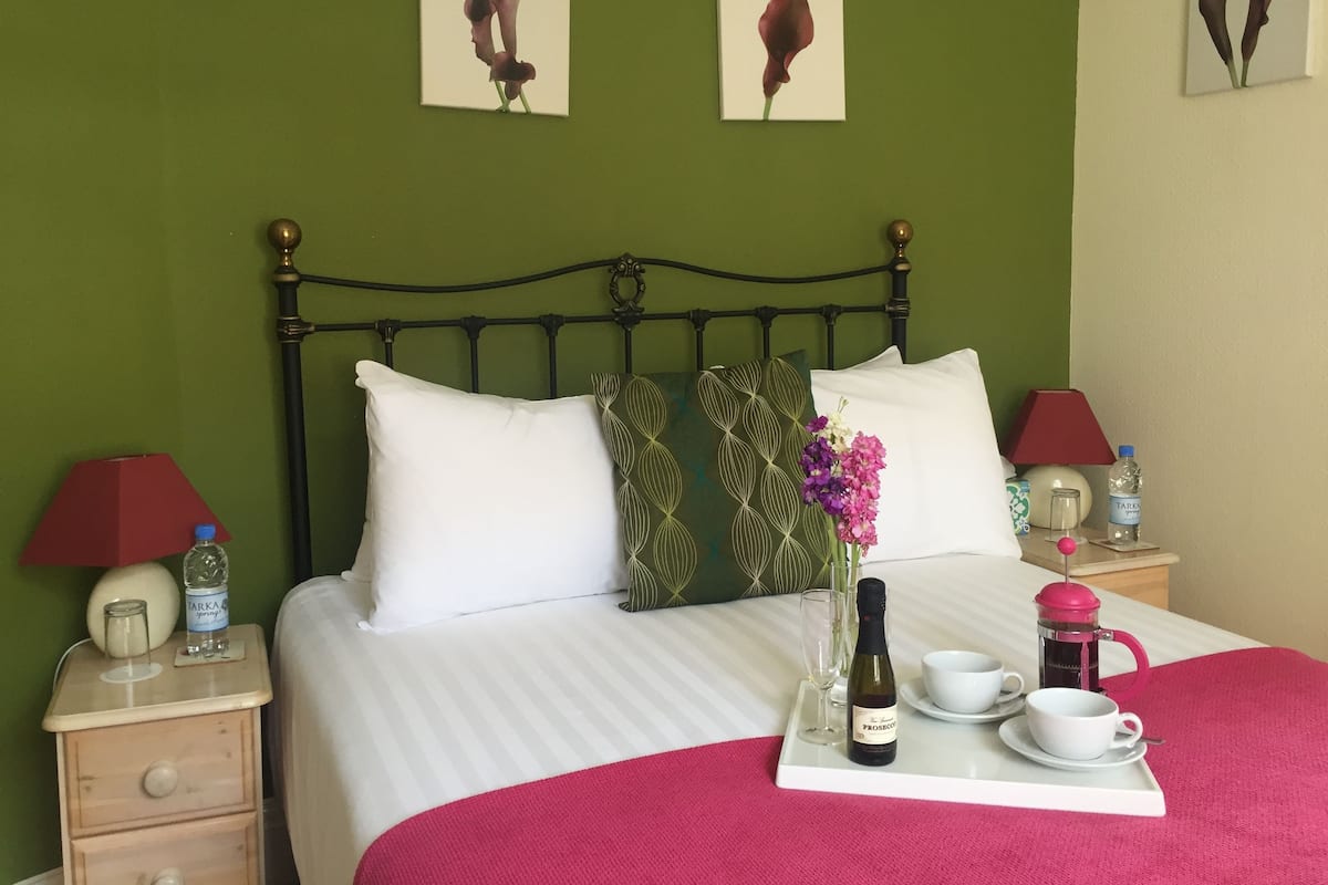 A neatly made bed with white linens, a red throw blanket, and decorative pillows. There is a bedside table with a red lamp, a water bottle, and a glass. A tray with a bottle of wine, a cup, and a sugar bowl is placed on the bed.