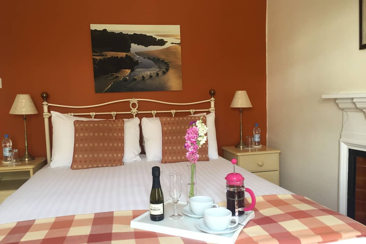 A neatly made bed with a checkered bedspread, a bedside table with a lamp, a bottle of wine, two cups, and a coffee maker on a tray.
