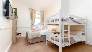 A bedroom with a bunk bed, a single bed, a TV mounted on the wall, and a window with curtains. - The Hollingworth (Lytham St. Anne's)