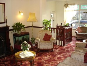 A traditional room with a fireplace, a wooden chair, a round table with a vase, and a view of the street through a window. - The Hollingworth (Lytham St. Anne's)