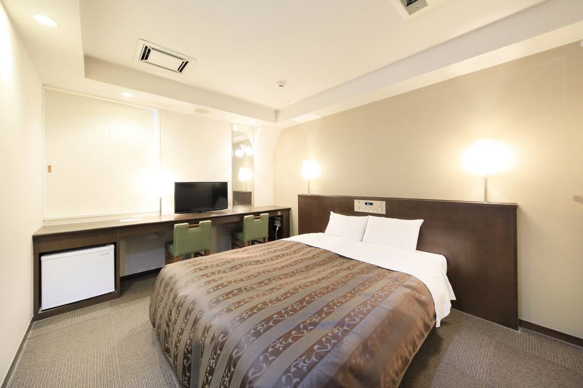 A hotel room with a bed, a desk with a TV, two chairs, and a mirror.