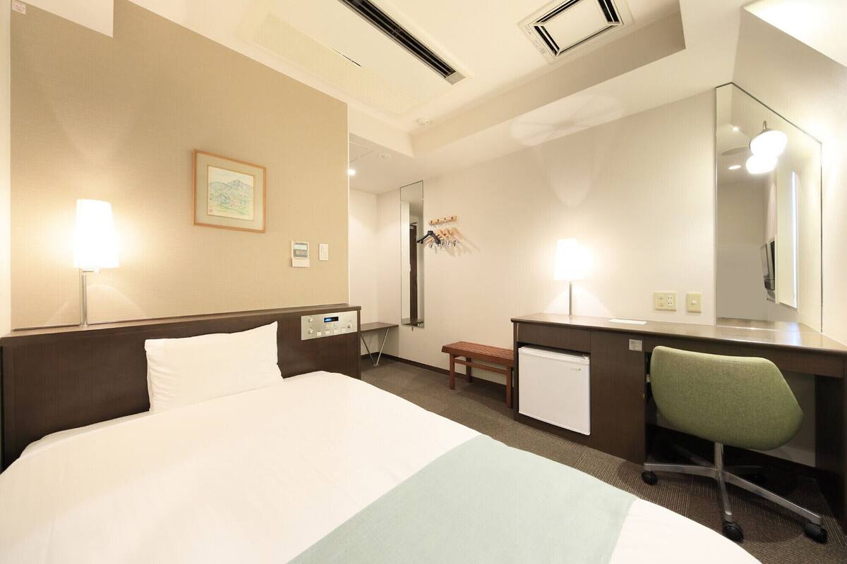 A hotel room with a bed, bedside lamps, a desk with a chair, a mirror, and a small bench.