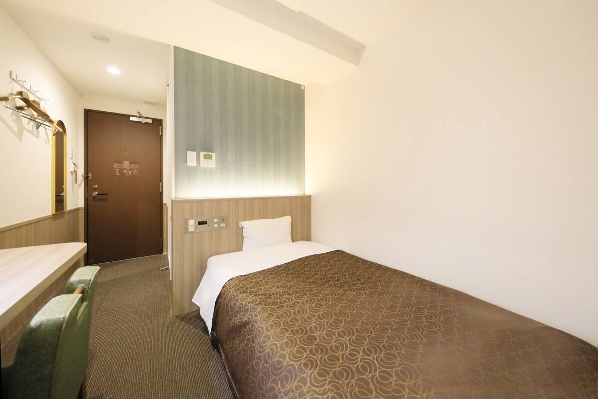 A hotel room with a bed, a bedside table, a mirror, a wall-mounted control panel, and a door.