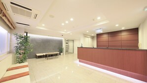 A reception area with a wooden reception desk, a bench, a plant, and a textured wall. - Hotel Shinjukuya (Machida)