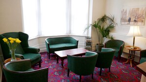 Quarto com cadeiras estofadas em verde, uma mesa de madeira, um vaso com flores amarelas, uma planta em vaso e um quadro na parede.