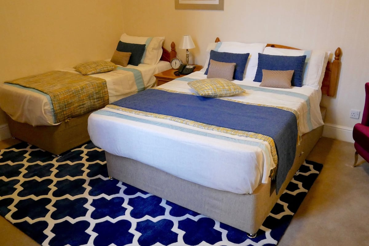 A hotel room with two beds, a red armchair, a patterned rug, and a bedside table with a lamp and phone.