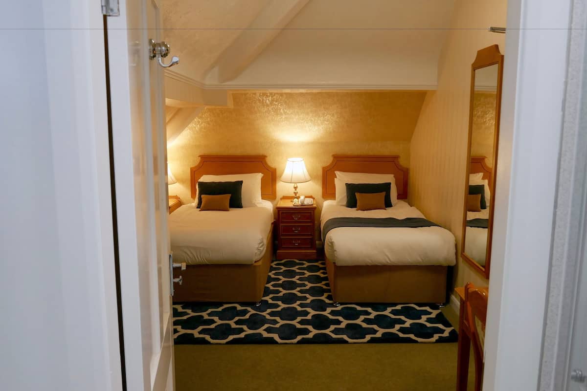 A hotel room with two beds, a nightstand with a lamp, a mirror, and a patterned carpet.