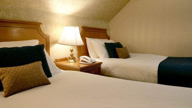 Quarto de hotel com duas camas, uma mesa de cabeceira com lâmpada e telefone, e uma cabeceira de madeira.