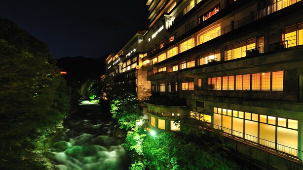 A multi-story hotel with illuminated windows and a river flowing through the courtyard. - Zazan Minakami (Minakami)