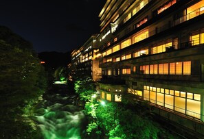 A multi-story hotel with illuminated windows and a river flowing through the courtyard. - Zazan Minakami (Minakami)