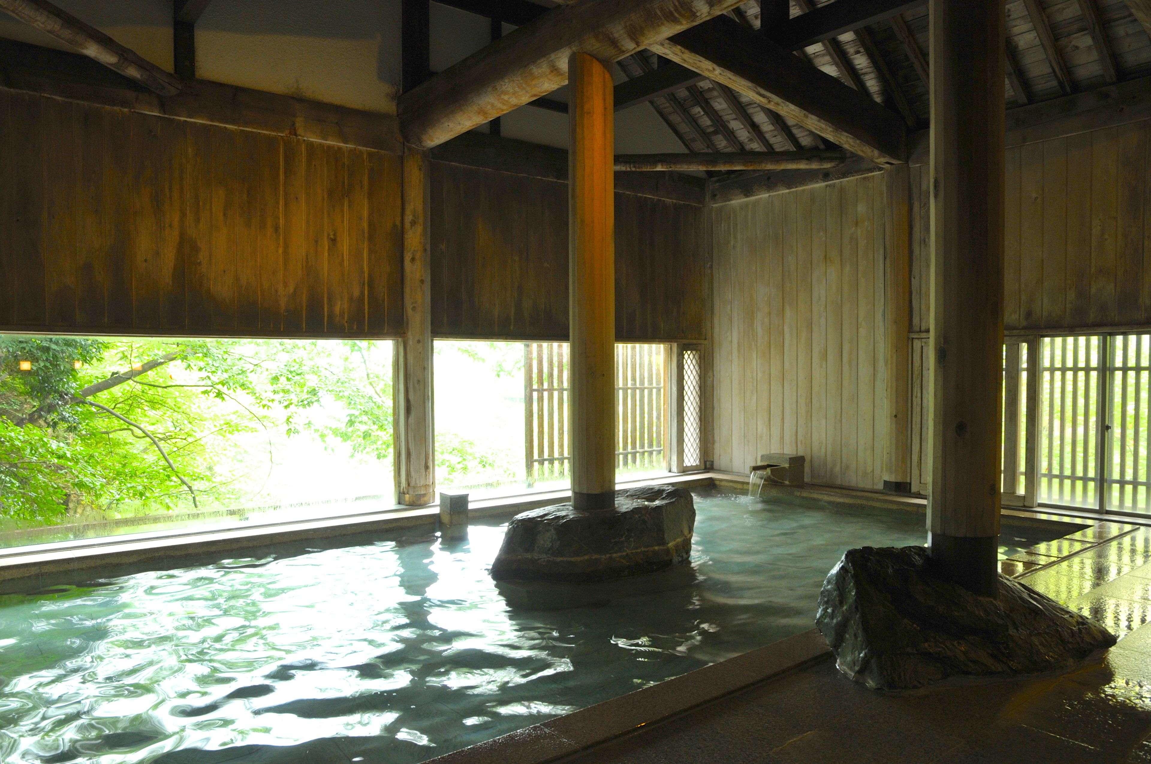 A traditional wooden bath with stone pillars, large windows, and a view of greenery outside.