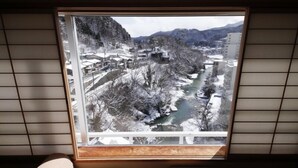A room with a large window offering a view of a snow-covered landscape with a river and buildings. - Zazan Minakami (Minakami)