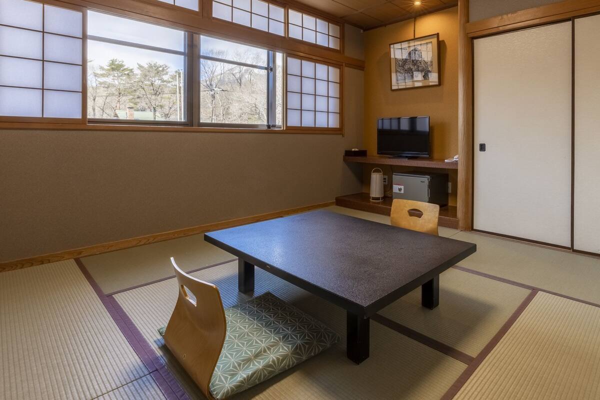 Una habitación japonesa tradicional con suelo de tatami, una mesa baja, un banco acolchado, un televisor y una puerta corredera.