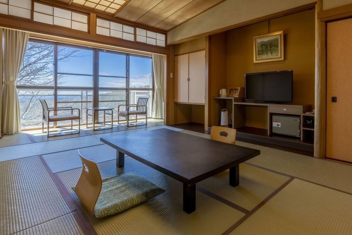 Una habitación japonesa tradicional con suelo de tatami, una mesa baja, sillas, un televisor y vistas al exterior.