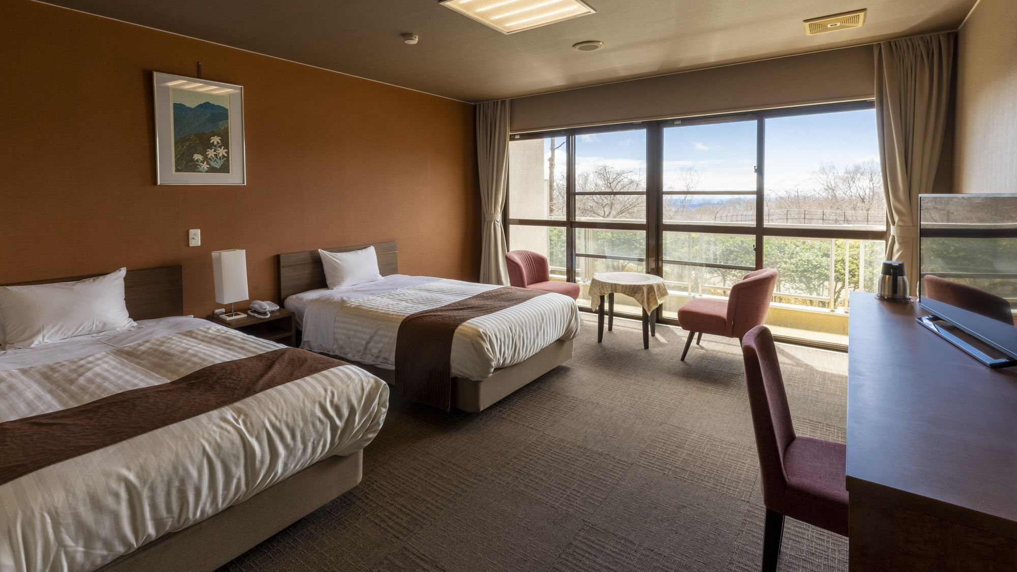 A hotel room with two beds, a desk, a chair, a small table, and a large window offering a view.