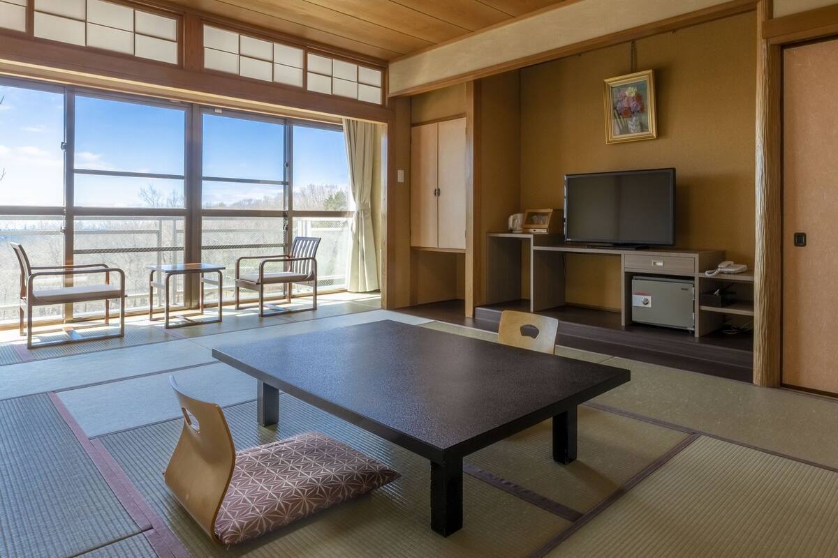 Una habitación tradicional japonesa con suelo de tatami, una mesa baja, sillas, un televisor, una caja fuerte y una puerta corredera.