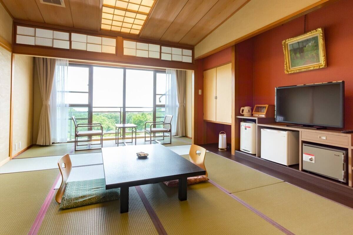 Una habitación japonesa tradicional con suelo de tatami, una mesita baja, sillas, un televisor y vistas a zonas verdes.