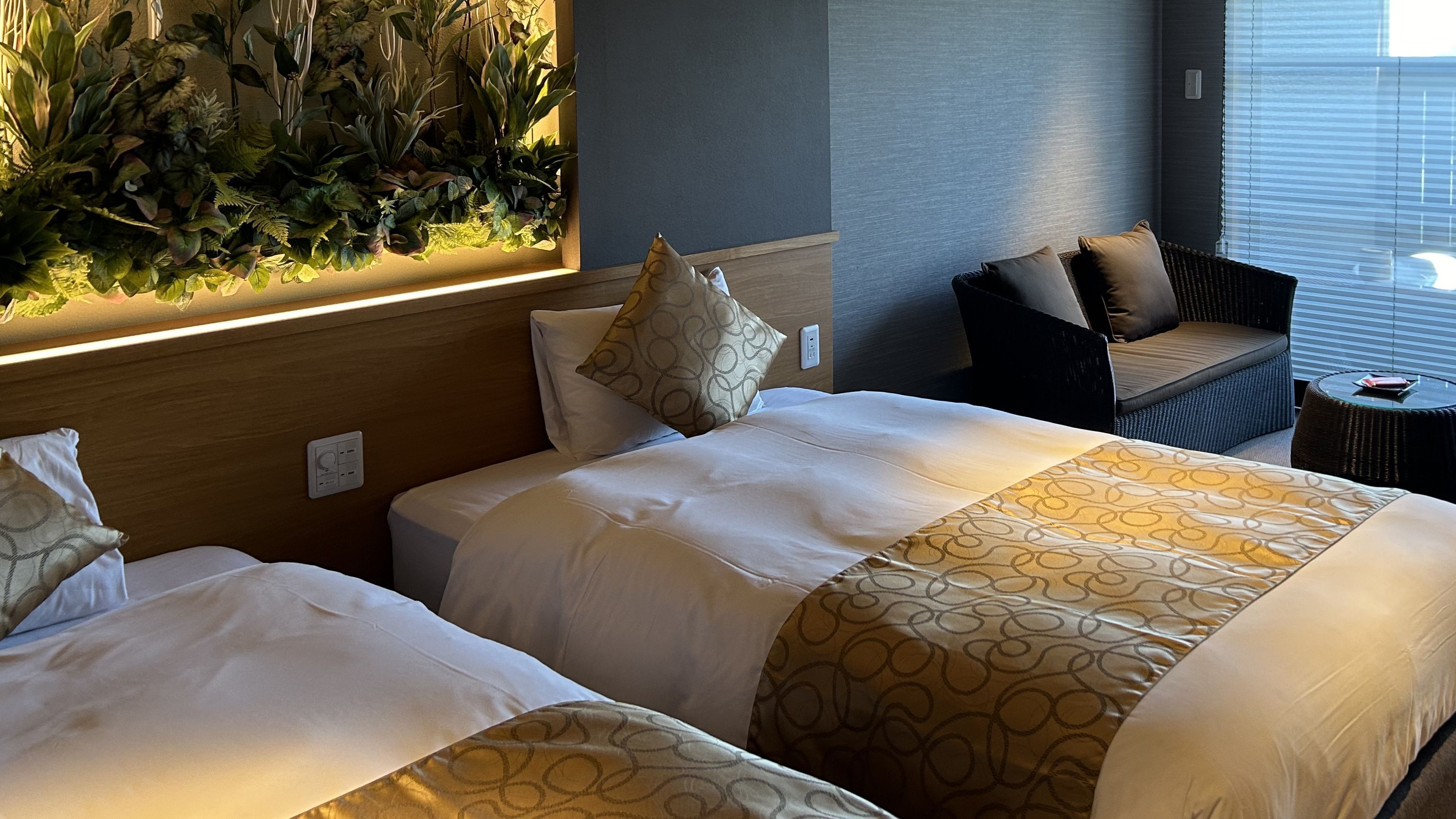 A hotel room with two beds, a wall-mounted plant, a seating area with a chair and ottoman, and a window with blinds.