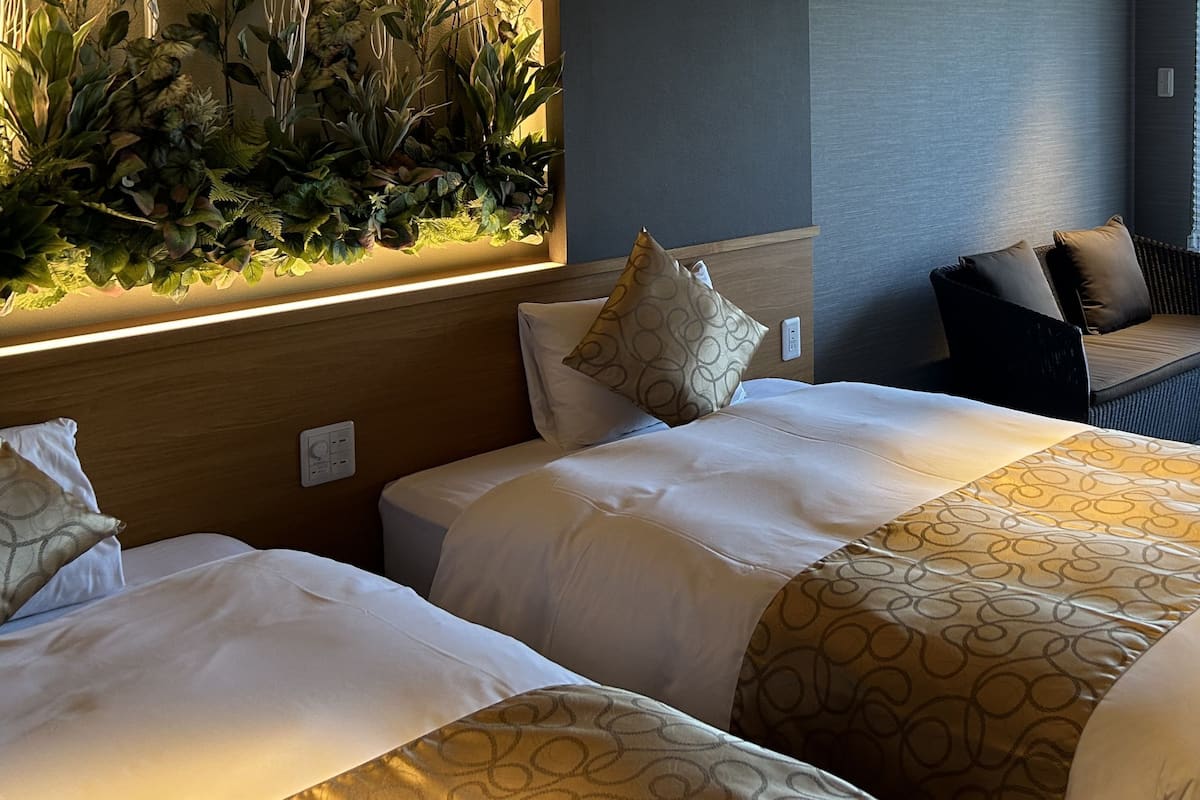 Habitación de hotel con dos camas, una planta en la pared, una zona de estar con un sillón y reposapiés, y una ventana con persianas.