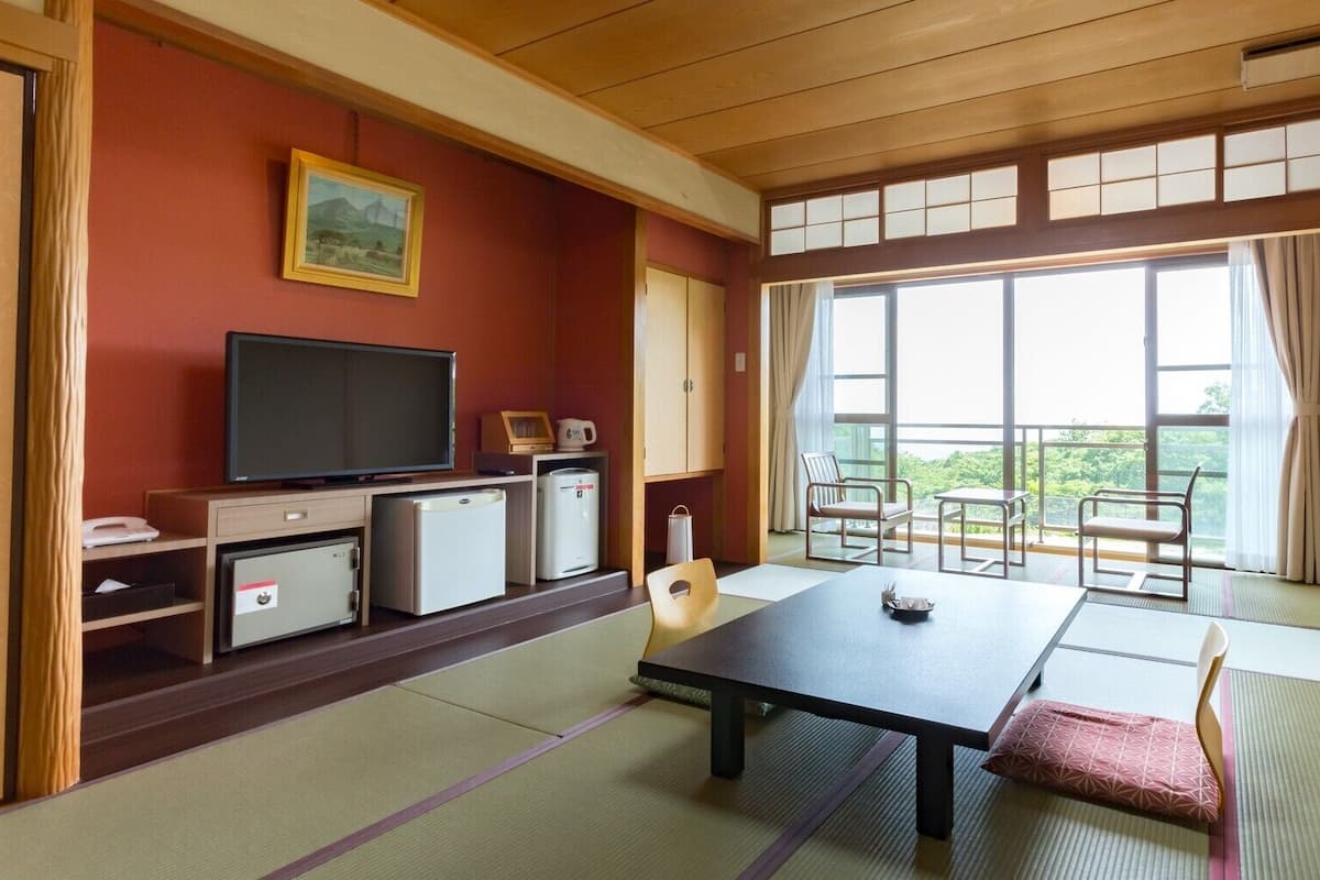 Una habitación japonesa tradicional con suelo de tatami, una mesa baja, un televisor y vistas a zonas verdes a través de amplias ventanas.