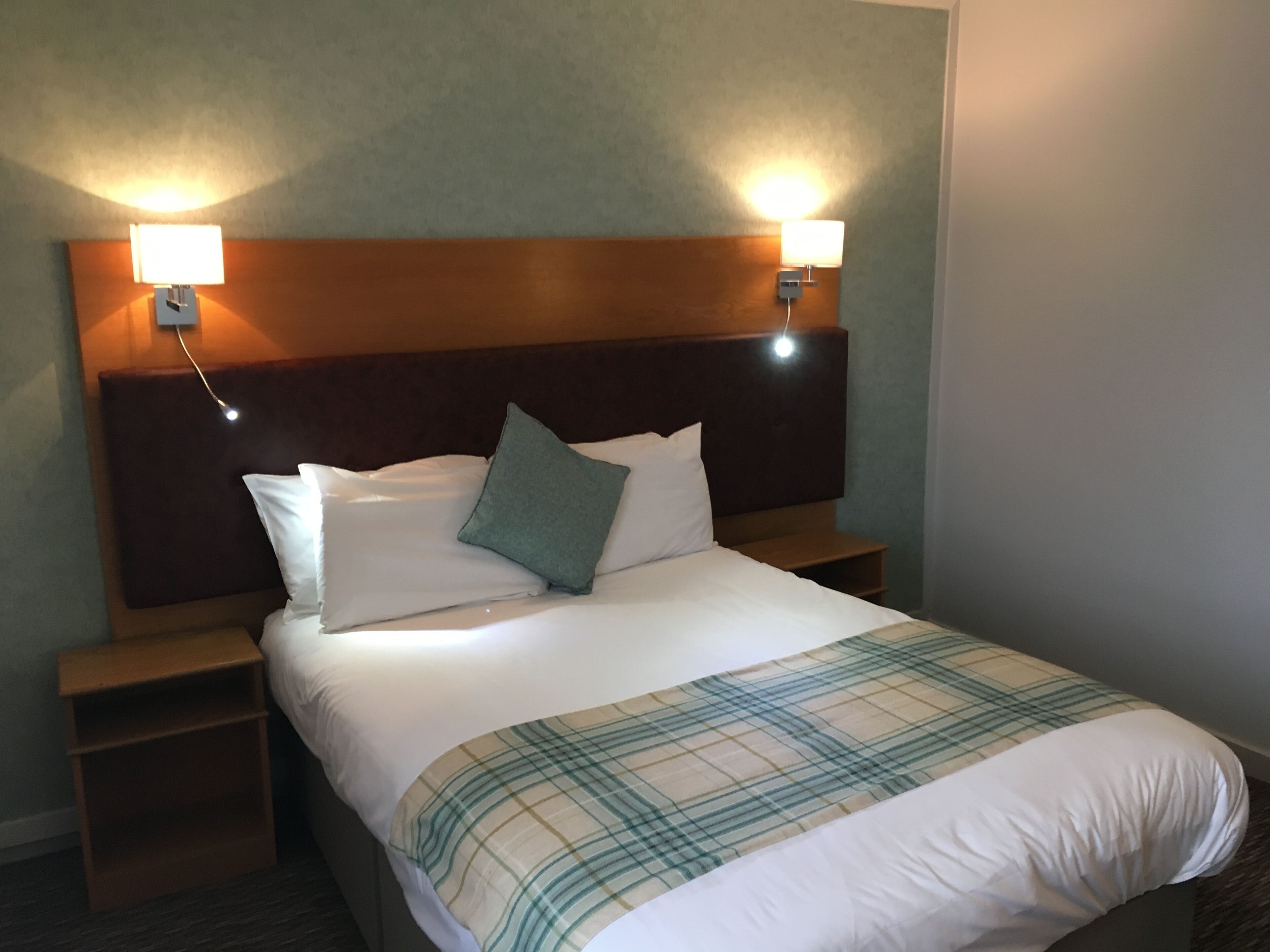 Una camera d'albergo con un letto, due comodini, un poggiabibite, una testiera e due lampade a parete.