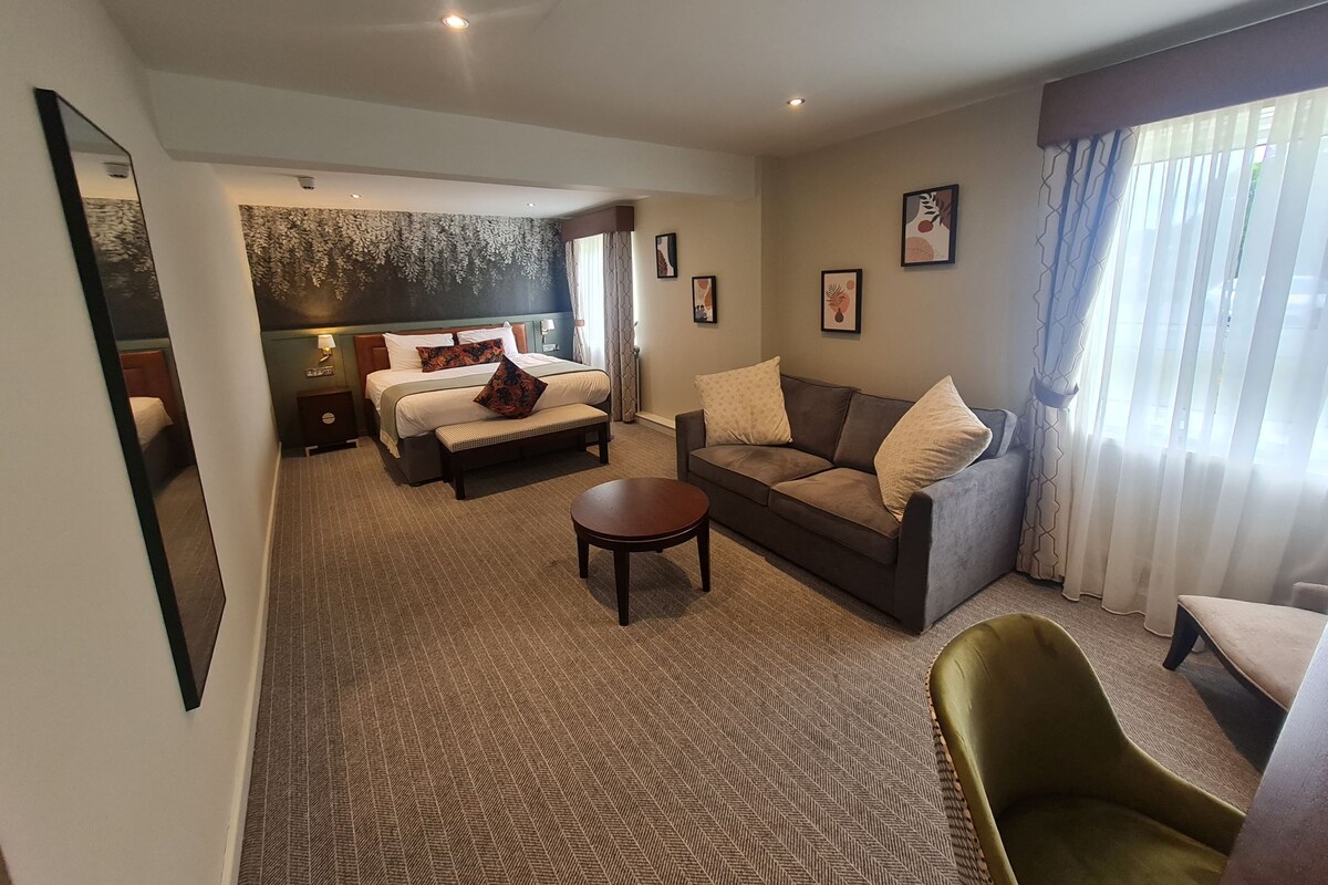 A hotel room with a bed, a sofa, a small table, a chair, and a large window with curtains.