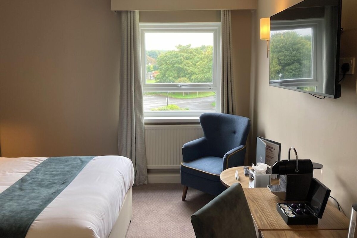 A hotel room with a bed, a desk with a chair, a window with curtains, and a flat-screen TV mounted on the wall.